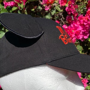 May include: A black baseball cap featuring two upright, rounded fabric ears attached to the top. The side of the cap displays embroidered text in bright orange thread that reads "My Birthday". The cap has a stiff, curved brim and a structured crown.