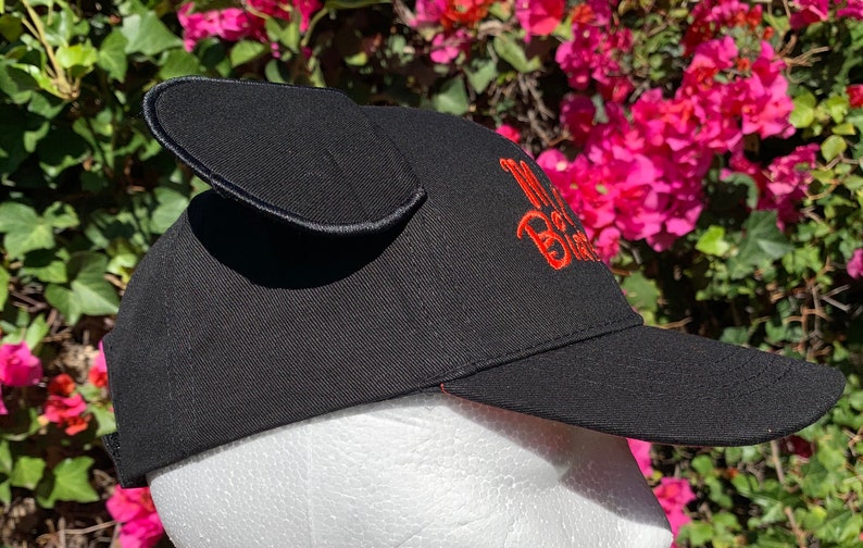 May include: A black baseball cap featuring two upright, rounded fabric ears attached to the top. The side of the cap displays embroidered text in bright orange thread that reads "My Birthday". The cap has a stiff, curved brim and a structured crown.