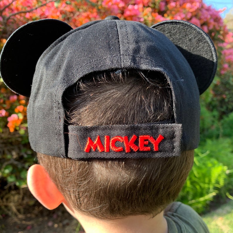 May include: A black baseball cap featuring two rounded black ears on the top and an adjustable strap at the back. The strap is embroidered with the word MICKEY in bold red capital letters, providing a clear view of the rear design details.
