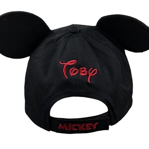 May include: A black baseball cap featuring two large, rounded black ears attached to the top. The name Toby is embroidered in red thread on the front, and the word MICKEY is embroidered in red thread on the adjustable back strap of the cap.