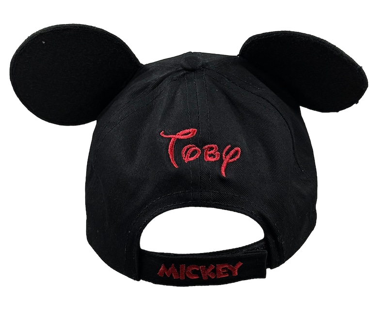 May include: A black baseball cap featuring two large, rounded black ears attached to the top. The name Toby is embroidered in red thread on the front, and the word MICKEY is embroidered in red thread on the adjustable back strap of the cap.