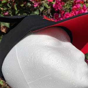 May include: A black baseball cap with a red underside on the brim and a small black protrusion on the top. The cap features orange embroidered text on the front panel and is displayed on a white foam mannequin head against a floral background.