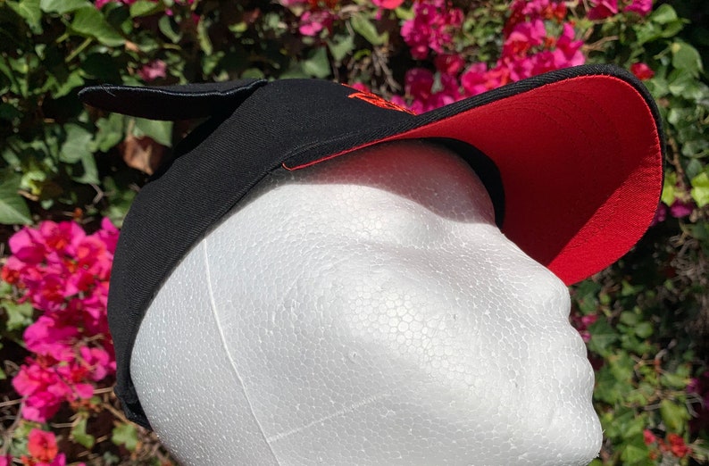 May include: A black baseball cap with a red underside on the brim and a small black protrusion on the top. The cap features orange embroidered text on the front panel and is displayed on a white foam mannequin head against a floral background.