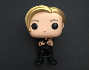 May include: A gold-haired pop culture figurine with black clothing and tattoos on its arms. The figurine is standing with its arms crossed.