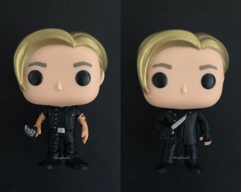 May include: A pop vinyl figure of a character with blond hair wearing a black leather jacket, black pants, and black boots. The figure is holding a knife in one hand and a small knife in the other. The figure is standing on a black background.