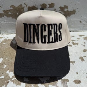 May include: A beige and black baseball cap with the word "DINGERS" embroidered in black block letters across the front. The cap has a structured crown and a curved brim.