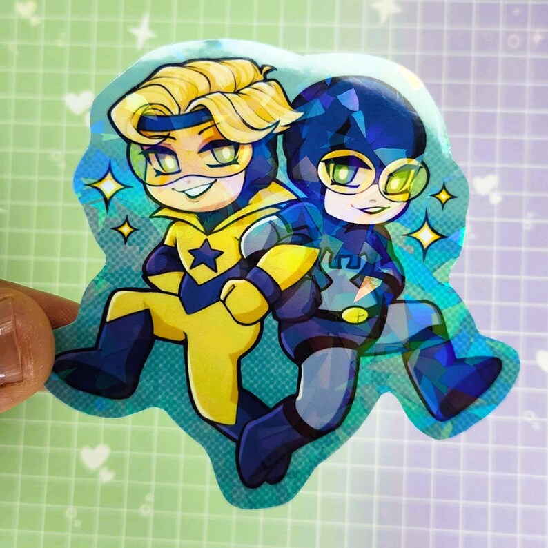 May include: A holographic sticker featuring two cartoon superheroes. One is in yellow and blue with a star emblem, the other in dark blue. Both have masks and are smiling, surrounded by sparkling stars.