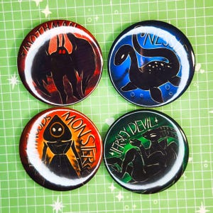 May include: Four circular badges featuring cryptid illustrations. The designs include the Mothman, Nessie, a figure labelled "Woods Monster", and the Jersey Devil. Each badge has a distinct colour scheme and design.