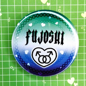 Puede incluir: Un bot&oacute;n redondo con un degradado azul y verde, con la palabra "FUJOSHI" en fuente g&oacute;tica negra. Un coraz&oacute;n blanco con dos s&iacute;mbolos masculinos en la parte inferior. El bot&oacute;n est&aacute; decorado con corazones blancos y un borde de cuentas.