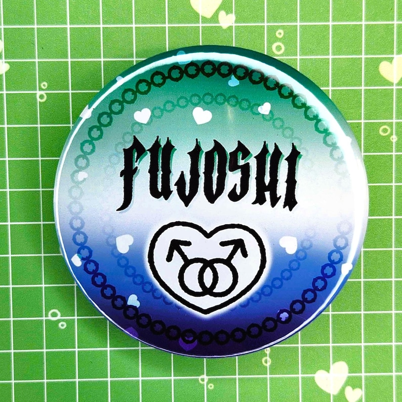 Puede incluir: Un bot&oacute;n redondo con un degradado azul y verde, con la palabra "FUJOSHI" en fuente g&oacute;tica negra. Un coraz&oacute;n blanco con dos s&iacute;mbolos masculinos en la parte inferior. El bot&oacute;n est&aacute; decorado con corazones blancos y un borde de cuentas.
