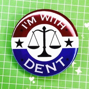 May include: A circular badge with a red and blue design. The badge features the text "I'M WITH DENT" in white, with a black scale of justice in the centre and black stars on either side.