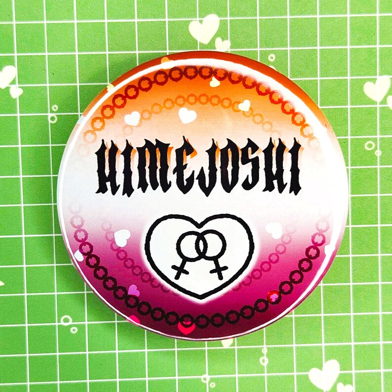 Puede incluir: Un bot&oacute;n redondo con un degradado de color de naranja a rosa. El bot&oacute;n presenta la palabra "HIMEJOSHI" en negro, un coraz&oacute;n con dos s&iacute;mbolos femeninos y un borde decorativo.