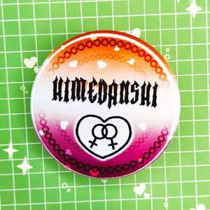 Puede incluir: Un bot&oacute;n redondo con un degradado de naranja, rosa y blanco. El bot&oacute;n presenta la palabra "HIMEDANSHI" en fuente g&oacute;tica negra, un coraz&oacute;n con dos s&iacute;mbolos femeninos y peque&ntilde;os corazones blancos.