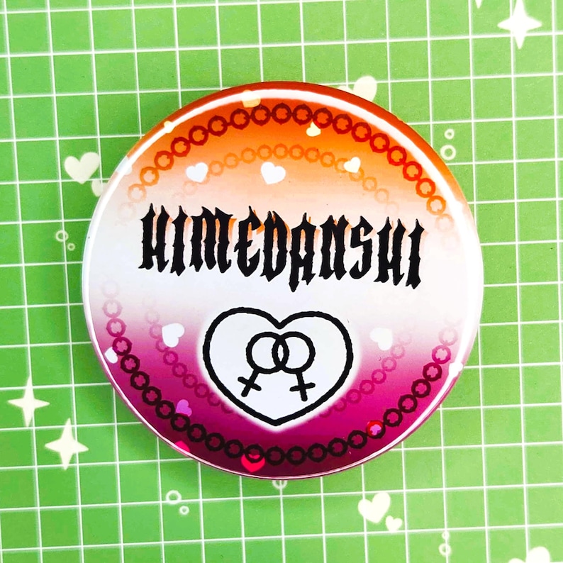 Puede incluir: Un bot&oacute;n redondo con un degradado de naranja, rosa y blanco. El bot&oacute;n presenta la palabra "HIMEDANSHI" en fuente g&oacute;tica negra, un coraz&oacute;n con dos s&iacute;mbolos femeninos y peque&ntilde;os corazones blancos.