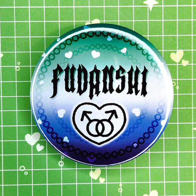Puede incluir: Un bot&oacute;n redondo con un degradado de azul y verde, con la palabra "FUDANSHI" en fuente g&oacute;tica negra. Un coraz&oacute;n blanco con dos s&iacute;mbolos masculinos en la parte inferior. El bot&oacute;n est&aacute; decorado con corazones blancos y cuentas azules.