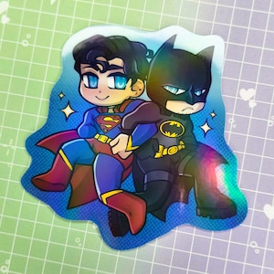 May include: A holographic sticker featuring cartoon versions of Superman and Batman. Superman is in a blue suit with a red cape, and Batman is in a black suit with a yellow bat symbol. Both characters are smiling and surrounded by star accents.