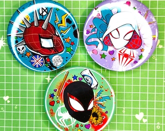 May include: Three round buttons featuring Spider-Man characters. One button has a red and blue Spider-Man design with spikes, another has a white and red Spider-Gwen design, and the third has a black and red Miles Morales design. The buttons are on a green grid background.