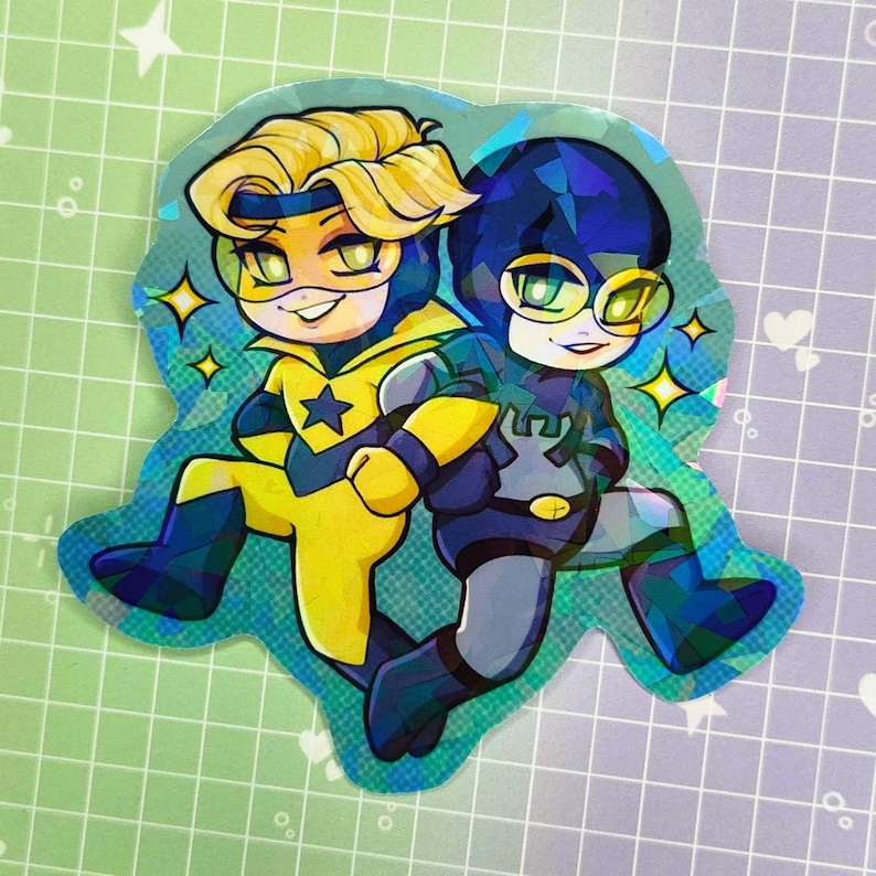 May include: A holographic sticker featuring two cartoon characters. One character is in yellow and blue with a star emblem, the other in blue and gray. Both characters are smiling and wearing masks. The sticker has a sparkly, iridescent finish.