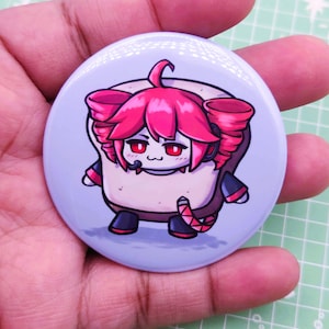 May include: A circular pin-back button featuring a cartoon illustration of a character with bright pink hair, red eyes, and a small mouth, set against a light purple background. The character has a square-shaped body and wears dark clothing with red accents.