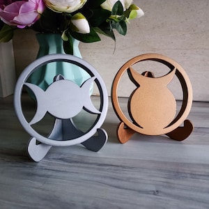 May include: Two circular decorative stands, one in a matte silver finish and one in a matte bronze finish. Each stand features a central cutout design of a triple moon symbol enclosed within a circular frame, supported by two small feet.
