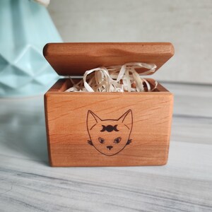 May include: A small, open wooden box with a cat face design. The box is made of light brown wood and contains shredded paper. The cat face has a crescent moon design above its eyes.