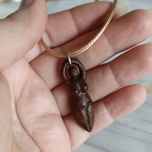 May include: A wooden pendant necklace featuring a carved figure of a woman with arms raised. The pendant is a dark brown color and is suspended from a light brown braided cord.