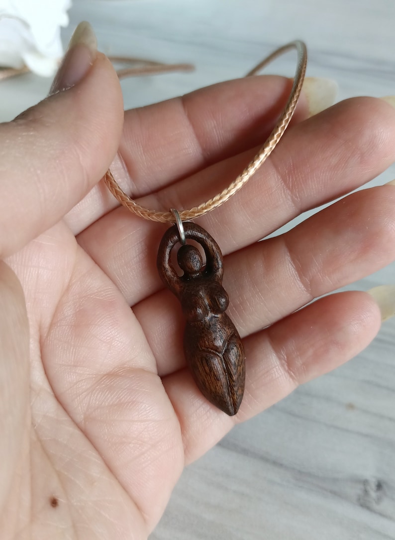 May include: A wooden pendant necklace featuring a carved figure of a woman with arms raised. The pendant is a dark brown color and is suspended from a light brown braided cord.