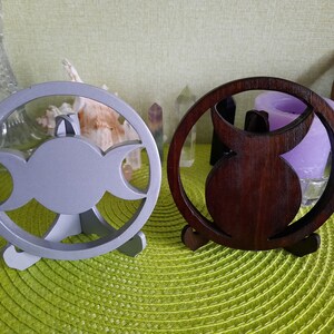 May include: Two circular wooden stands featuring a cutout triple moon symbol. One stand is painted a matte light gray, while the other has a dark brown wood grain finish. Each piece is supported by a small, integrated tripod base for stability.