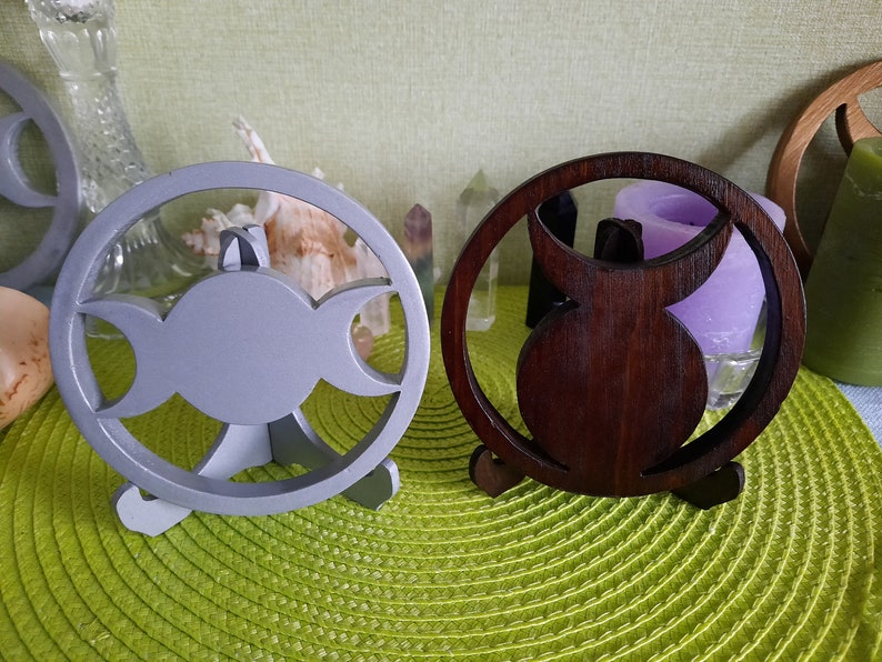 May include: Two circular wooden stands featuring a cutout triple moon symbol. One stand is painted a matte light gray, while the other has a dark brown wood grain finish. Each piece is supported by a small, integrated tripod base for stability.