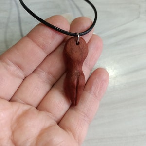 May include: A reddish-brown wooden pendant necklace featuring a carved abstract figure. The pendant is suspended from a black cord. The necklace is held in a hand.