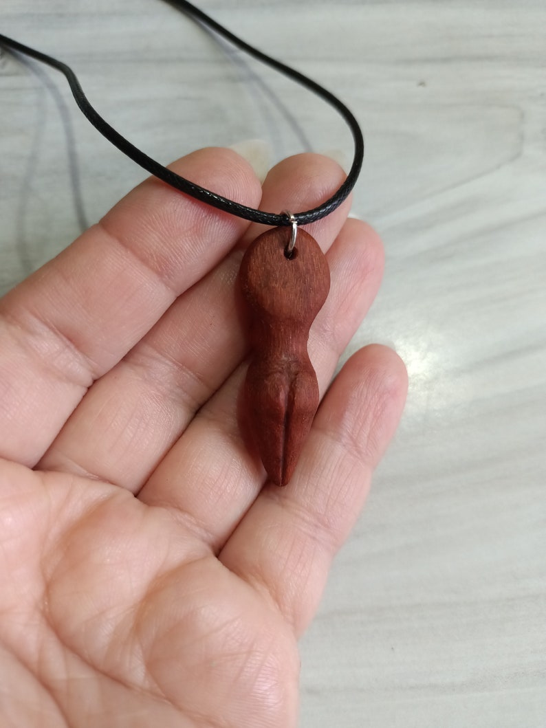 May include: A reddish-brown wooden pendant necklace featuring a carved abstract figure. The pendant is suspended from a black cord. The necklace is held in a hand.