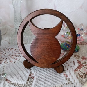 May include: A circular dark brown wooden stand featuring a central cutout design that resembles a crescent moon shape. The object is supported by two small, curved wooden feet at the base, allowing it to stand upright on a flat surface.