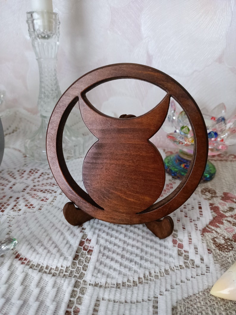 May include: A circular dark brown wooden stand featuring a central cutout design that resembles a crescent moon shape. The object is supported by two small, curved wooden feet at the base, allowing it to stand upright on a flat surface.