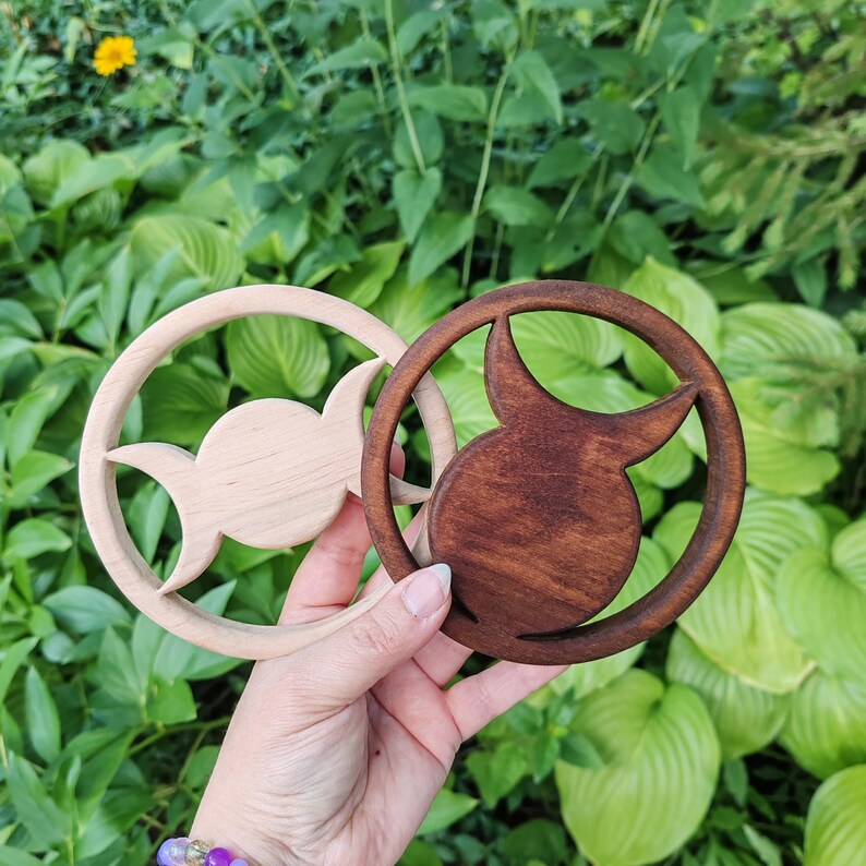 May include: Two circular wooden ornaments featuring a cutout triple moon symbol. One ornament is crafted from light-colored natural wood, while the other is finished in a dark brown stain. Both pieces have a smooth, rounded frame design.