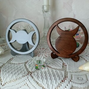 May include: Two circular decorative stands, one in a matte silver finish and one in a dark brown wood finish. Each stand features a central cutout design representing a triple moon symbol, supported by a small base for upright display.