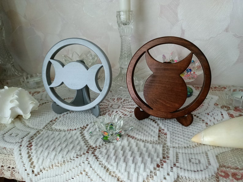 May include: Two circular decorative stands, one in a matte silver finish and one in a dark brown wood finish. Each stand features a central cutout design representing a triple moon symbol, supported by a small base for upright display.
