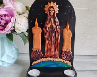 Può includere: Un altare in legno del Giorno dei Morti con una figura scolpita con un teschio, mani giunte in preghiera e un'aureola. Due sculture a forma di candela affiancano la figura. L'altare ha una base nera con due portacandele e un disegno della terra dipinto.