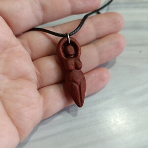 May include: A carved wooden pendant depicting a stylized female figure. The pendant is a rich reddish-brown color and is suspended from a black cord. The figure has a rounded head, arms raised above the head, and a defined torso.
