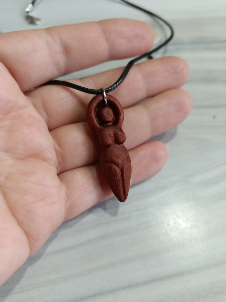 May include: A carved wooden pendant depicting a stylized female figure. The pendant is a rich reddish-brown color and is suspended from a black cord. The figure has a rounded head, arms raised above the head, and a defined torso.