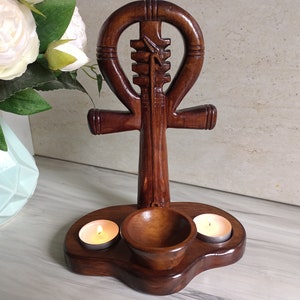 May include: A handcrafted wooden ankh candle holder. The dark brown ankh features a cross shape with a loop at the top and a small bowl and two candle holders at the base. Two lit tea light candles are in the holders.