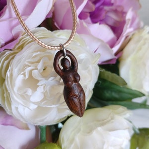 May include: A dark brown wooden pendant necklace featuring a carved female figure. The pendant is suspended from a beige cord. The background includes white and purple flowers, creating a soft, natural aesthetic.