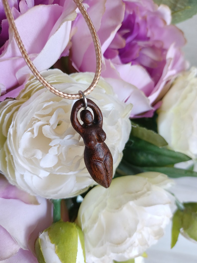 May include: A dark brown wooden pendant necklace featuring a carved female figure. The pendant is suspended from a beige cord. The background includes white and purple flowers, creating a soft, natural aesthetic.