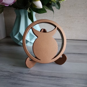 May include: A circular bronze-colored stand featuring a central silhouette of a horned figure within a ring frame. The object is supported by two curved feet at the base, providing a stable structure for display purposes on a flat surface.