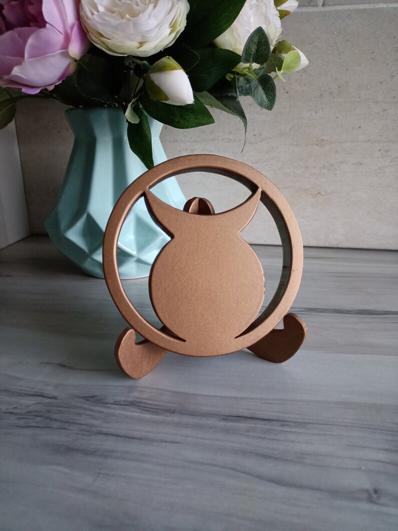 May include: A circular bronze-colored stand featuring a central silhouette of a horned figure within a ring frame. The object is supported by two curved feet at the base, providing a stable structure for display purposes on a flat surface.