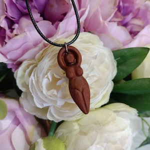May include: A wooden pendant necklace featuring a carved figure of a woman with arms raised. The pendant is a reddish-brown color and hangs from a black cord. The background is a soft focus of white and pink flowers.