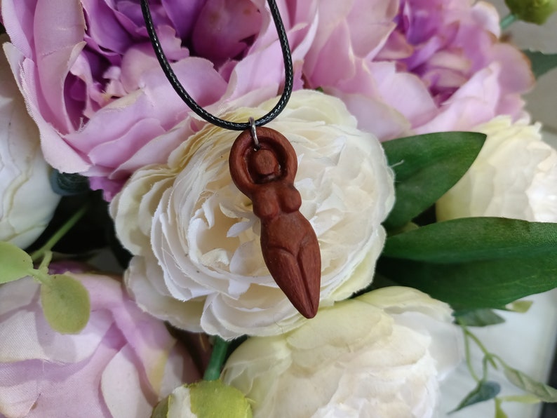 May include: A wooden pendant necklace featuring a carved figure of a woman with arms raised. The pendant is a reddish-brown color and hangs from a black cord. The background is a soft focus of white and pink flowers.
