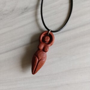 May include: A wooden pendant necklace featuring a carved figure of a woman. The pendant is a reddish-brown color and hangs from a black cord. The figure has a stylized form with rounded shapes.