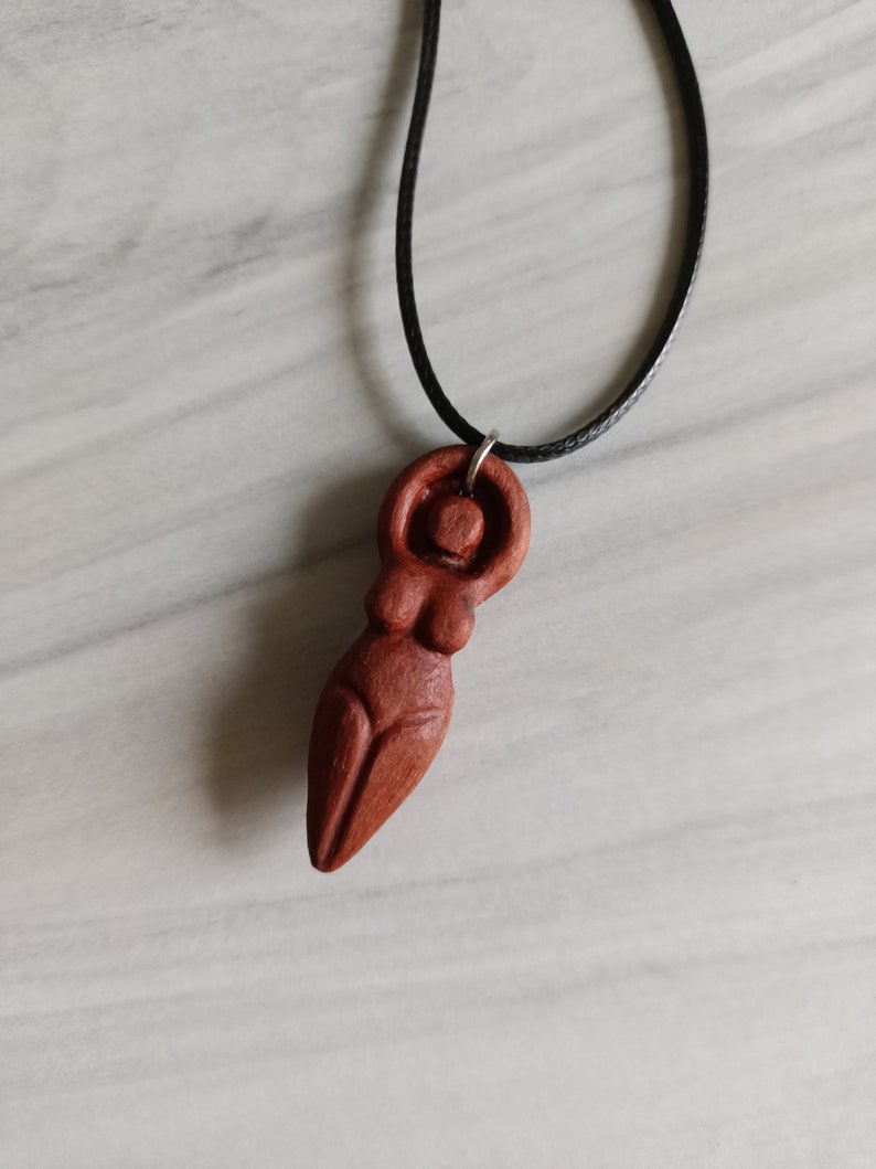 May include: A wooden pendant necklace featuring a carved figure of a woman. The pendant is a reddish-brown color and hangs from a black cord. The figure has a stylized form with rounded shapes.