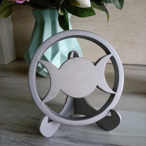 May include: A silver-colored circular stand featuring a triple moon symbol cutout design. The object is supported by a three-legged base, providing a stable structure for display. The finish is matte and uniform, highlighting the geometric cutouts.