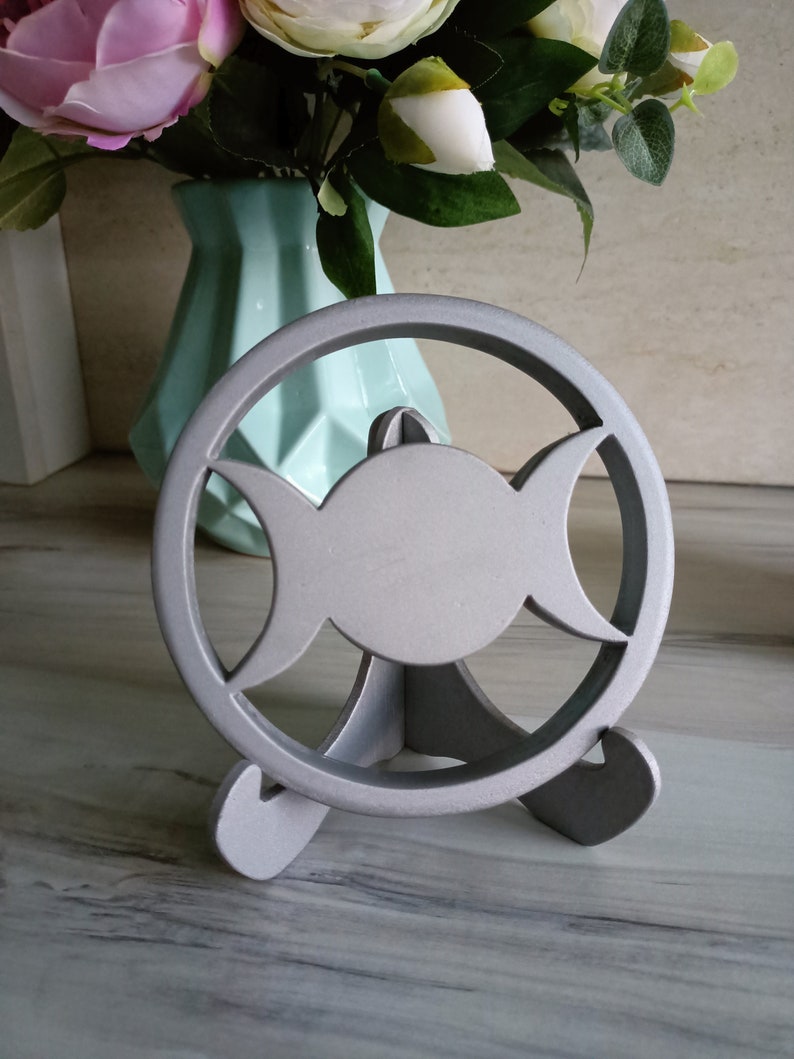 May include: A silver-colored circular stand featuring a triple moon symbol cutout design. The object is supported by a three-legged base, providing a stable structure for display. The finish is matte and uniform, highlighting the geometric cutouts.